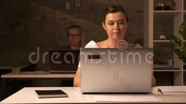 两个工作的白种人妇女坐在棕色的办公室里，在室内的白色办公桌上使用笔记本电脑